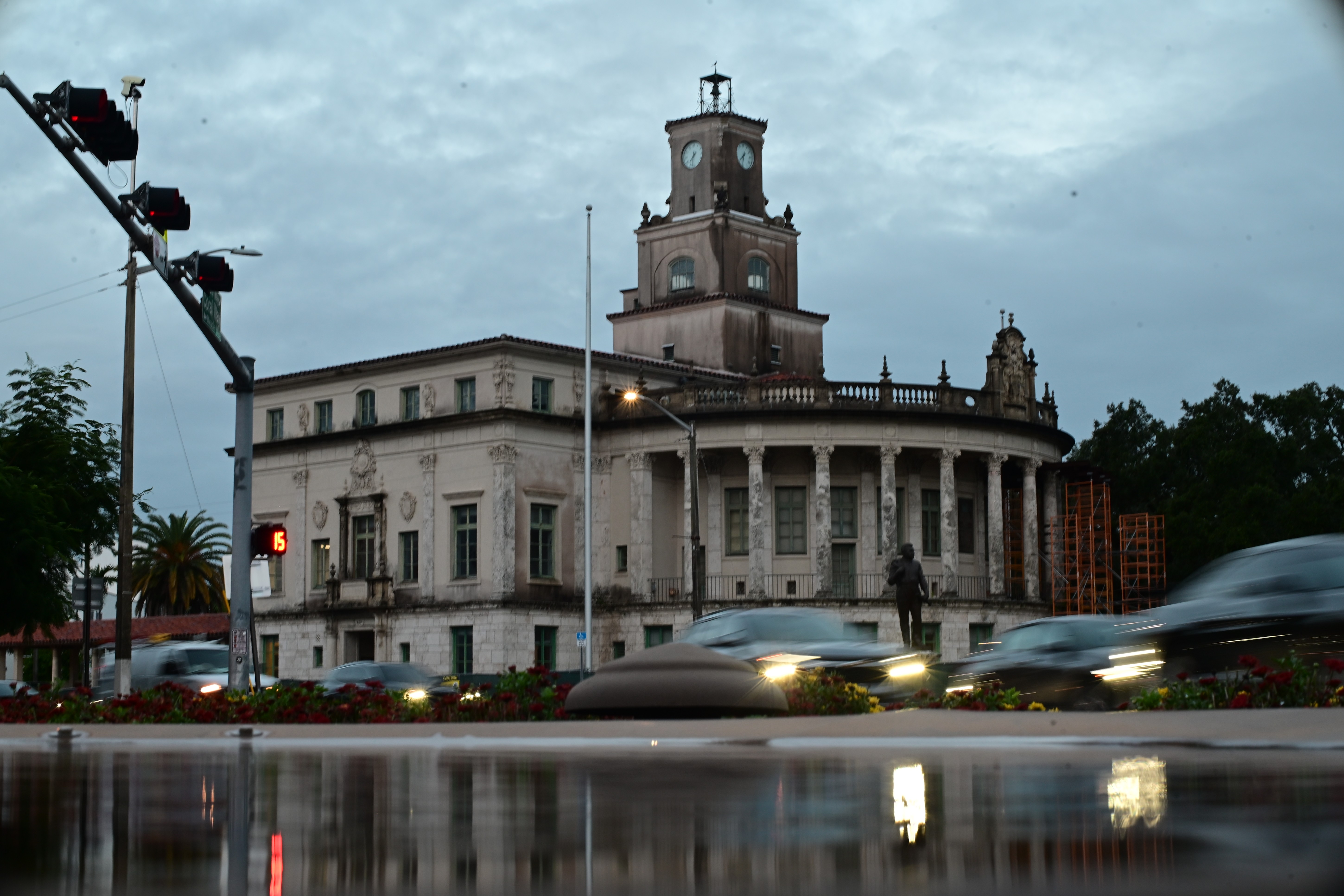 A clock tower beneath a cloudy sky, with passing cars and reflections in the foreground.