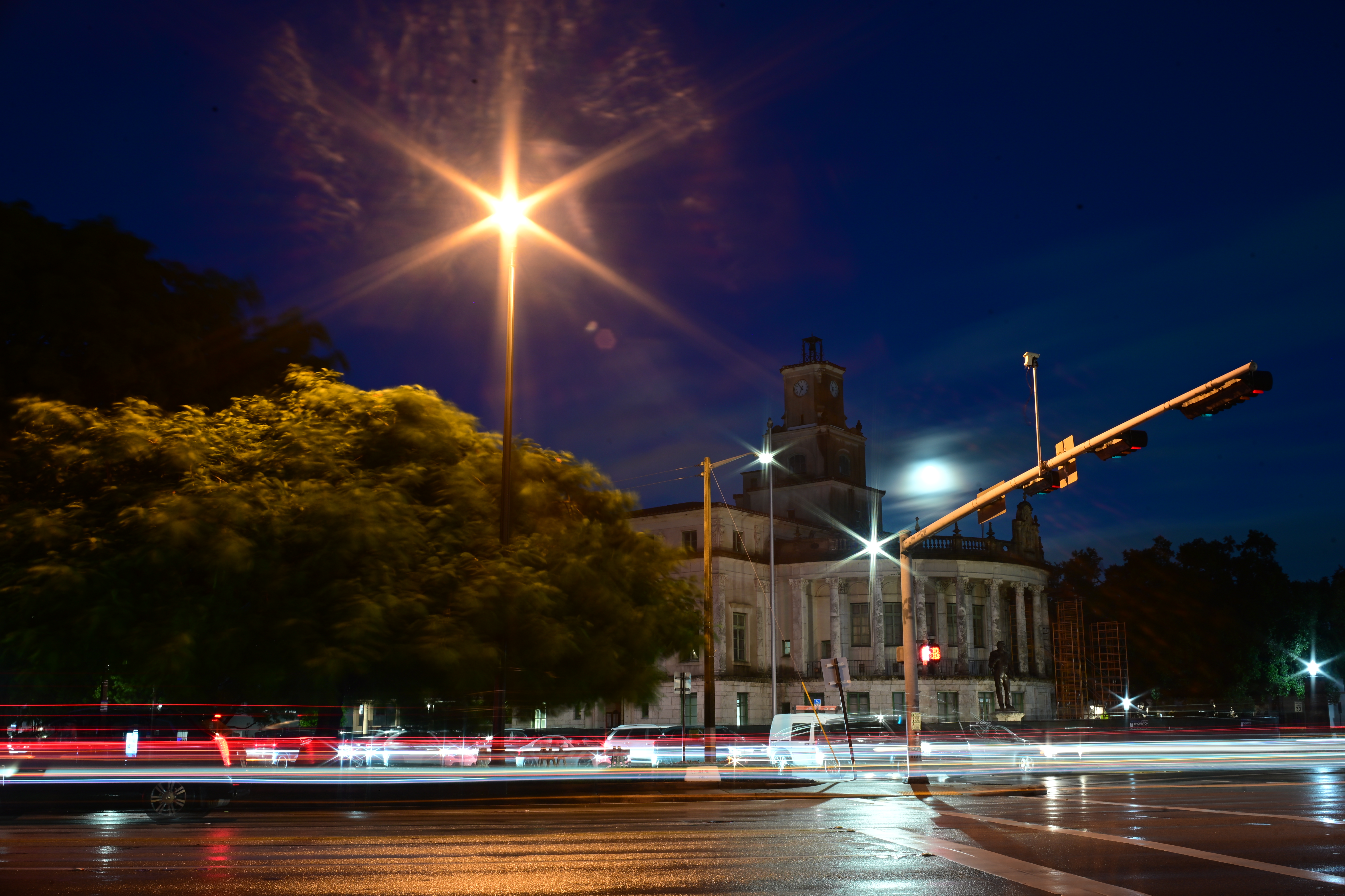 Light trails cross a rain-soaked street beneath a clock tower and a starburst streetlight.