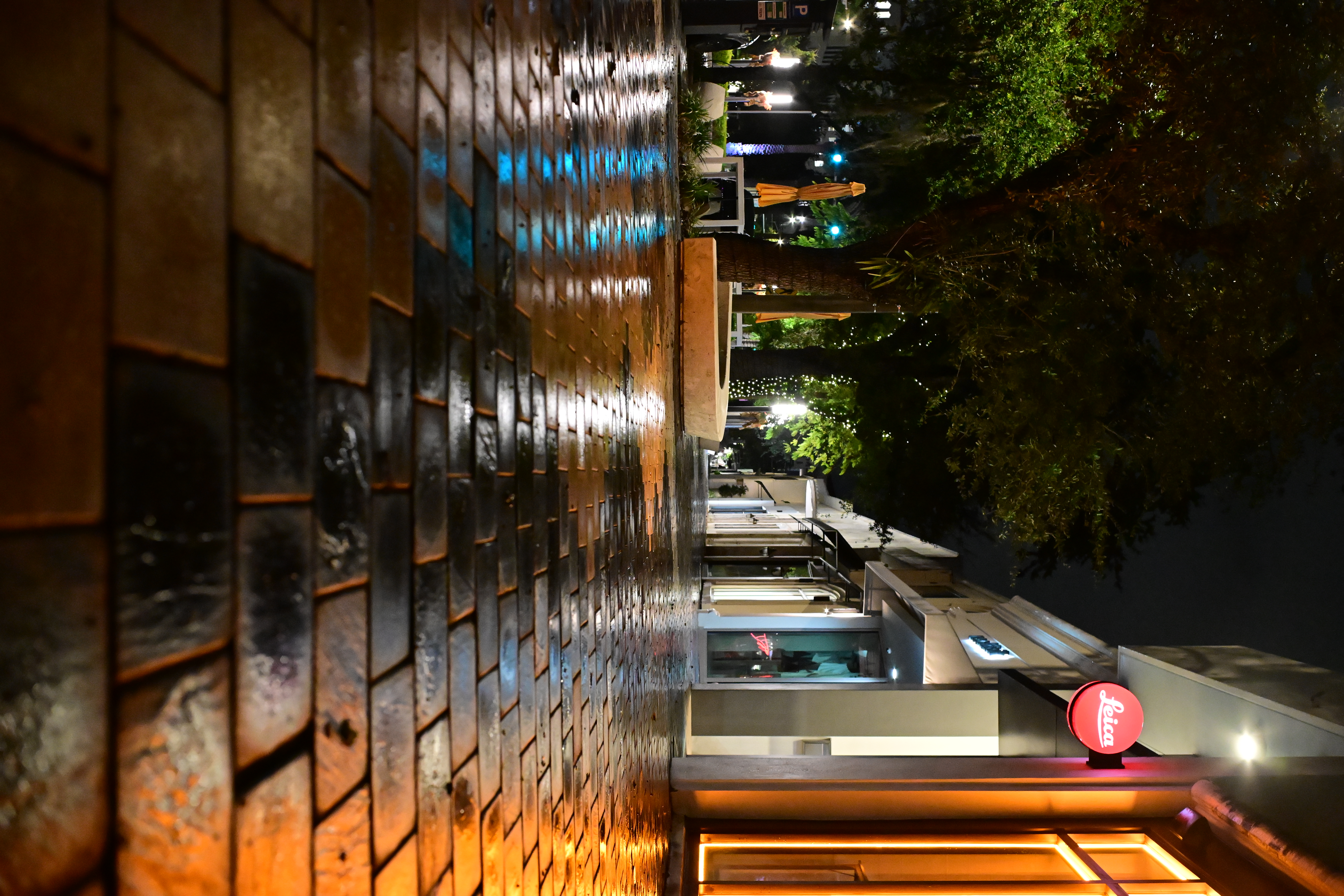 Amber shop windows and a red Leica sign reflected in wet paving stones at night.